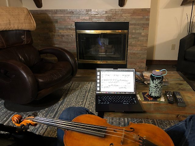 A violin resting in the foreground of a cozy living room, with a laptop showing sheet music set up for an online lesson.