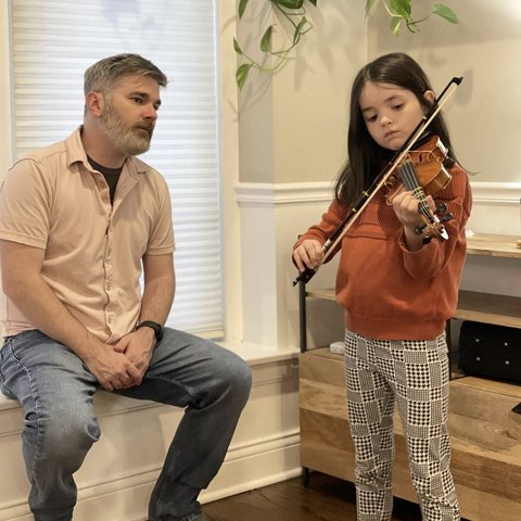 A father sits nearby as his young daughter practices violin at home.