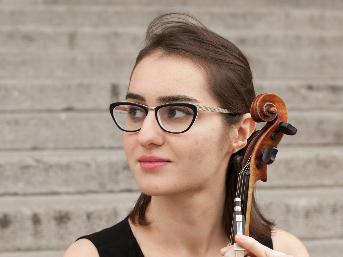 An adult violinist wearing glasses, holding her violin by the scroll and looking off to the side.