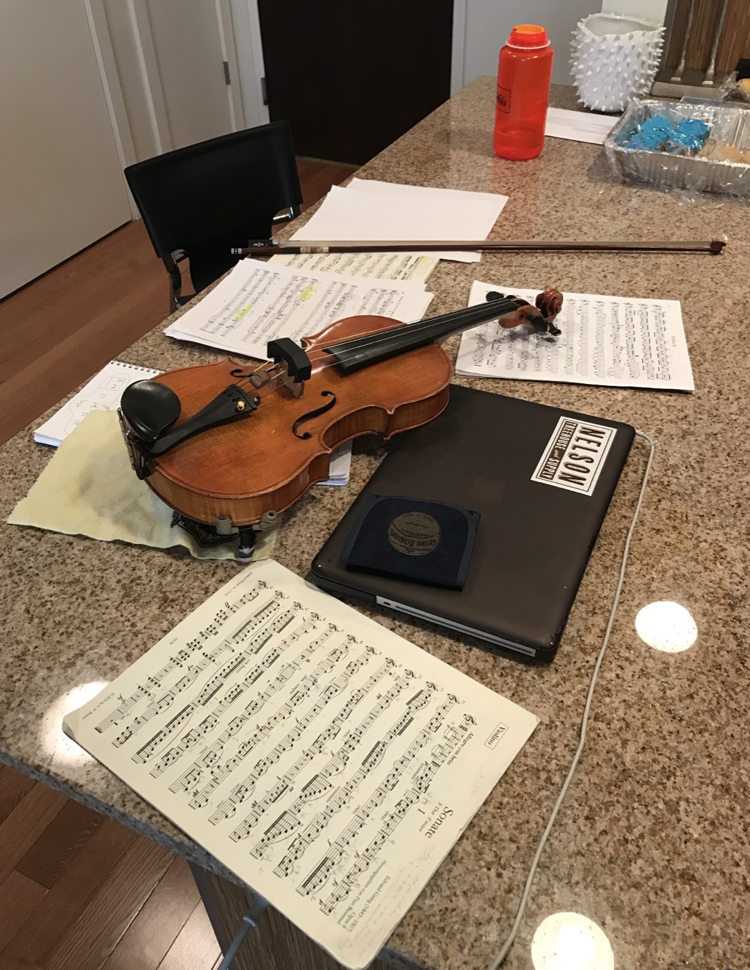 A violin resting on a kitchen counter beside its bow, open sheet music, and a laptop, set up for home practice.