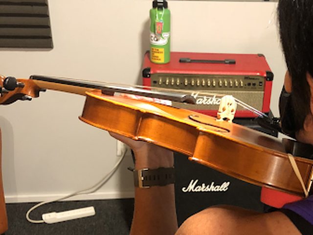A student practicing violin indoors, bow drawn across the strings, in a home studio setting.