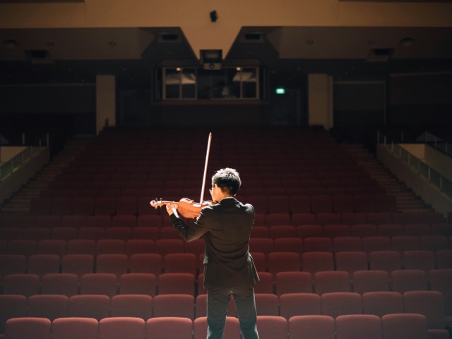 A young violinist in a suit stands alone on a stage, playing to an empty theater lined with red seats.