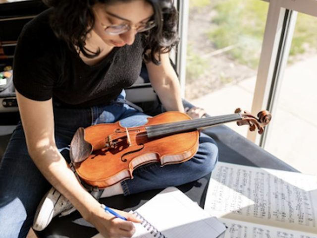 An adult student sitting by a sunny window, violin in her lap, writing notes in a practice journal next to sheet music.