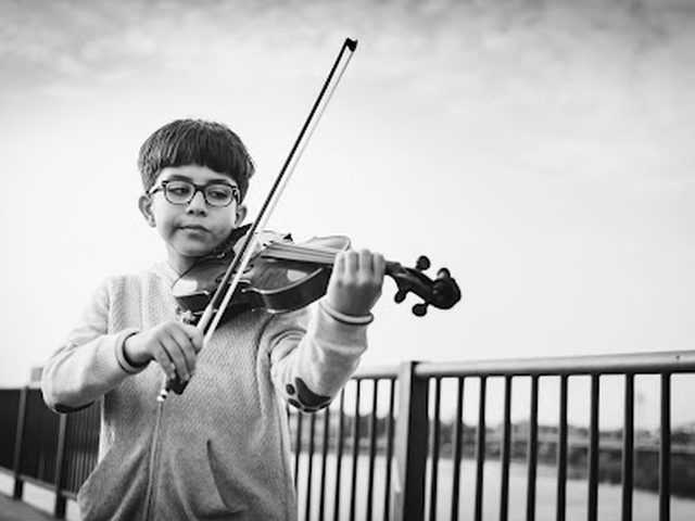 Black-and-white photo of a young boy playing violin outdoors on a bridge, focused on his bow hand.